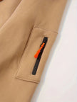 Close-up of a brown jacket with a zipper pocket featuring an orange zipper pull on a white background.