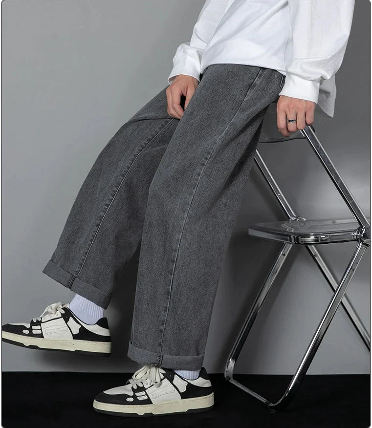 Person wearing gray wide-leg jeans and black and white sneakers sitting on a small metal stool against a plain background.