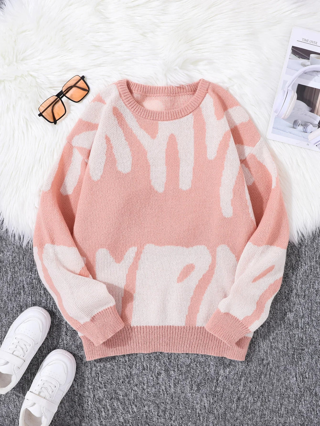 Pink sweater with white abstract patterns on a gray surface with white sneakers and sunglasses.