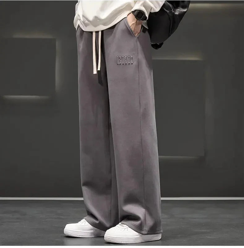 Person wearing gray wide-leg pants with a brand logo, white sneakers, and a white hoodie against a dark background.