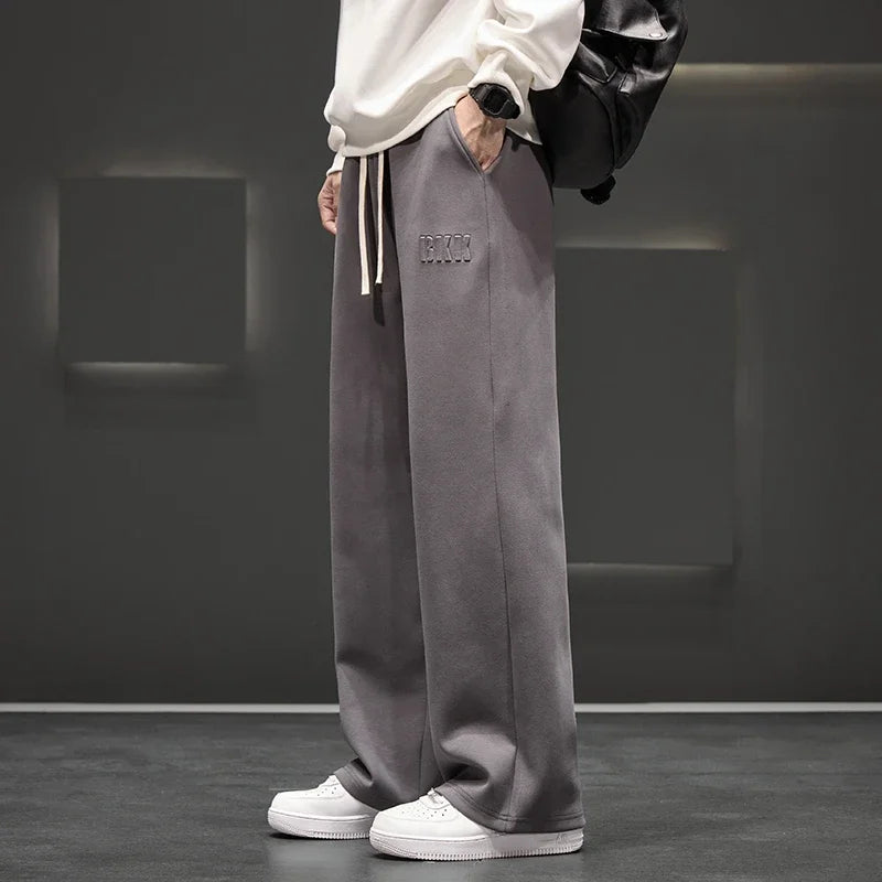 Person wearing gray wide-leg pants with a brand logo, white sneakers, and a black backpack against a dark background.