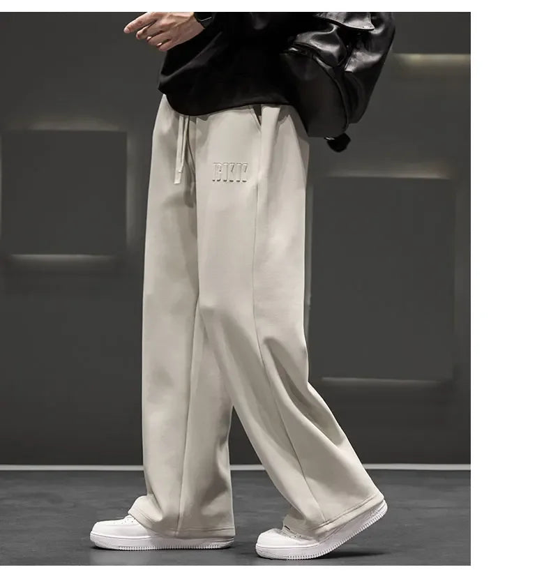 Person wearing beige high-waisted pants with a brand logo, standing against a dark background.