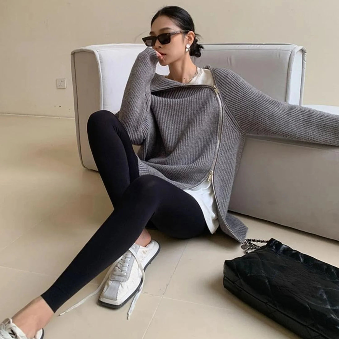 Woman sitting on a couch wearing a gray sweater and black leggings with a black handbag beside her.