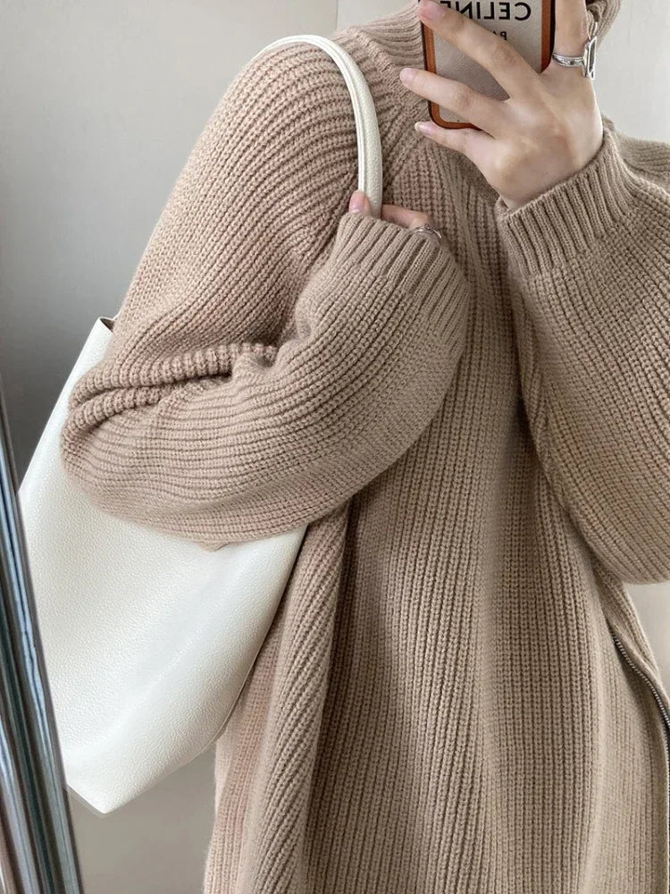 Person wearing a beige knitted sweater holding a phone with a white strap.