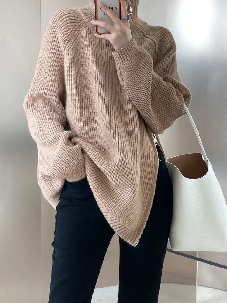 Person wearing a beige sweater and dark jeans, holding a phone with a neutral background.