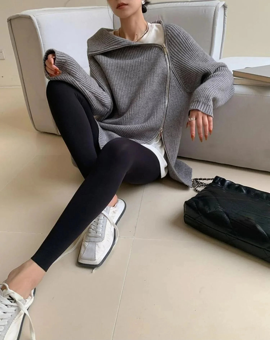 Person sitting on a couch wearing a gray sweater, black leggings, and white sneakers.